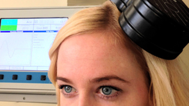 A woman sits with a device near her head and a computer screen in the background.