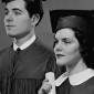 A black and white photo of a heterosexual couple wearing caps and gowns and holding diplomas.