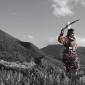 A Native woman wearing traditional Native clothing holds a large feather in the hair. She is standing on the side of a mountain or hill.