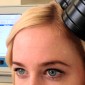 A woman sits with a device near her head and a computer screen in the background.