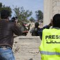 A photojournalist with a neon-yellow vest that says "Press" across the back uses a camera in a combat zone