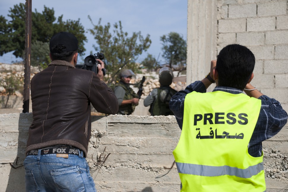 A photojournalist with a neon-yellow vest that says "Press" across the back uses a camera in a combat zone