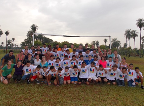 A group photo shows many young people, both boys and girls, wearing matching T-shirts and posing in front of a soccer goal.