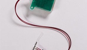 The elongated tab (near the tip of the tweezers) is the soft, stretchable sensor. The green box is the implantable "base station," which holds the electrical components to power the device and wirelessly transmit data.