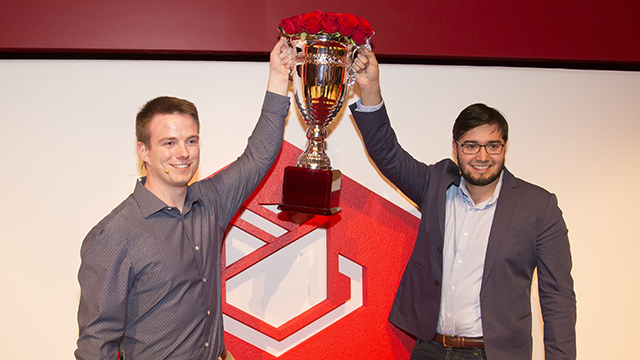 Two people hold a large trophy with roses inside.