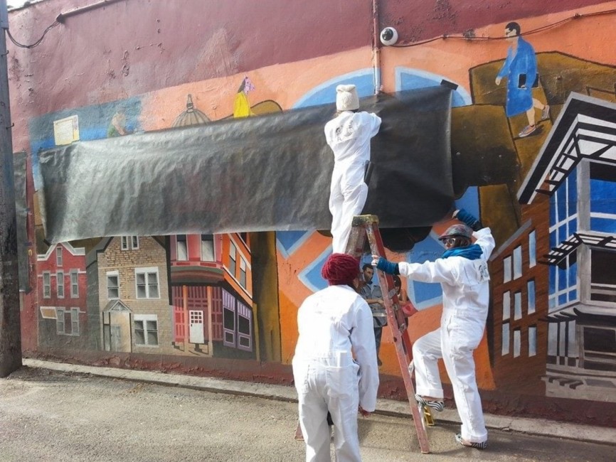 Three people wearing white painter suits cover up a mural with a tarp.