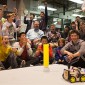 A group of people cheer while looking at a small robot vehicle on the floor.