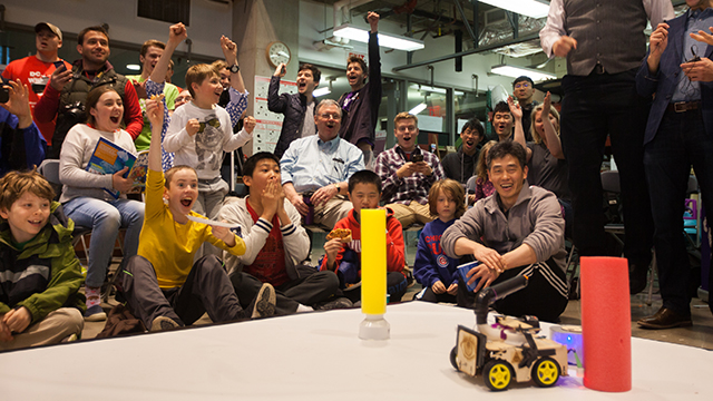 A group of people cheer while looking at a small robot vehicle on the floor.