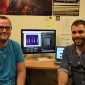 Two men sit in front of a computer.