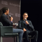 Trevor Noah holds a microphone and sits in a chair on a stage