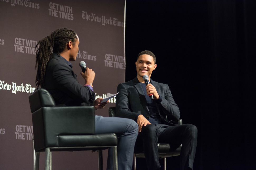 Trevor Noah holds a microphone and sits in a chair on a stage