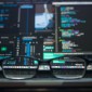 A pair of glasses sits on a table with several computer monitors behind it.