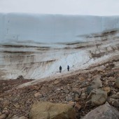 Two people stand in front of a tall ice sheet.