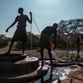 Two men collect water from a well. There are sevarl camels in the background.