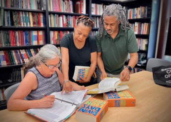 surrouned by bookshelves, three people look at a book manuscript and the novel "Divine Days" 