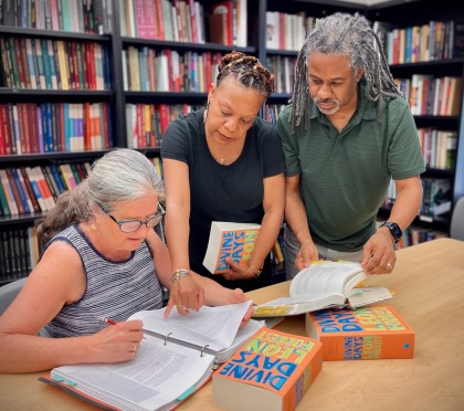 surrouned by bookshelves, three people look at a book manuscript and the novel "Divine Days" 