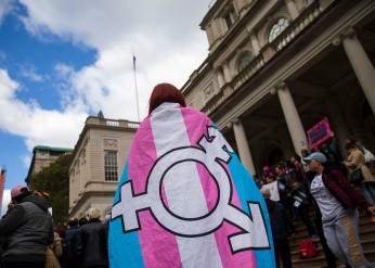 A person wearing a trans flag emblazoned with the gender inclusivity symbol