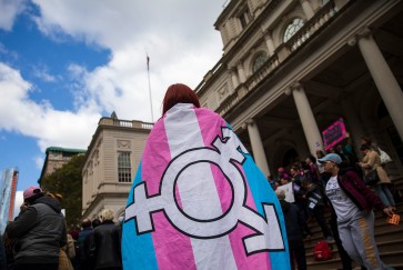 A person wearing a trans flag emblazoned with the gender inclusivity symbol