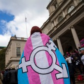 A person wearing a trans flag emblazoned with the gender inclusivity symbol