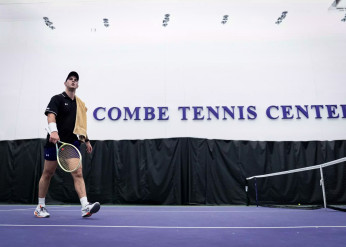 A men's tennis team member walks on a court with a Combe Tennis Center sign in the background