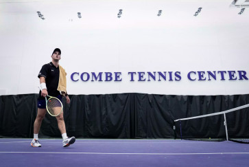 A men's tennis team member walks on a court with a Combe Tennis Center sign in the background
