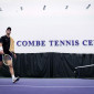 A men's tennis team member walks on a court with a Combe Tennis Center sign in the background