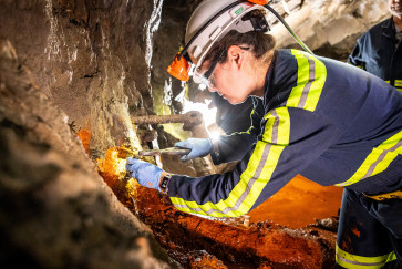 a woman in reflective gear and a hard hat chisels at a rock wall