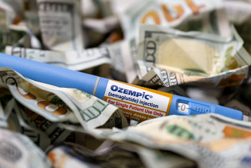 A blue Ozempic GLP-1 autoinjector sits in a pile of crumpled $100 bills