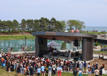 A crowd gathers in front of Kahn Pavilion, where a rock band plays, with the lagoon and Lake Michigan in the background