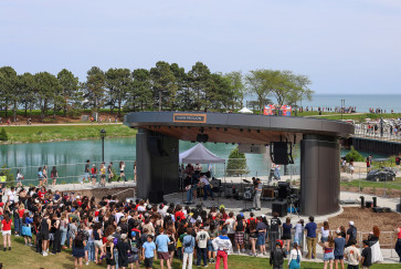A crowd gathers in front of Kahn Pavilion, where a rock band plays, with the lagoon and Lake Michigan in the background