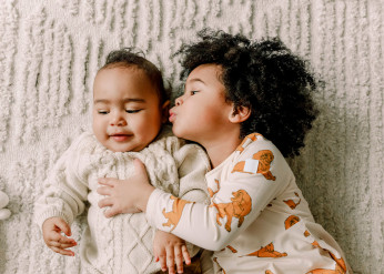 A baby lying on a blanket receives a kiss from an older sibling