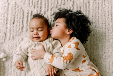 A baby lying on a blanket receives a kiss from an older sibling