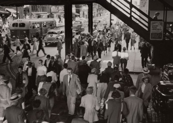 A black and white photo of pedestrians crossing a road under an elevated train overpass