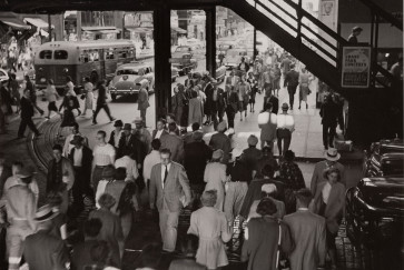 A black and white photo of pedestrians crossing a road under an elevated train overpass