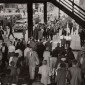 A black and white photo of pedestrians crossing a road under an elevated train overpass