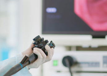 A gloved hands holds the controls of a scope for a colonscopy with a computer screen in the background
