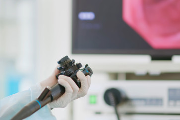 A gloved hands holds the controls of a scope for a colonscopy with a computer screen in the background