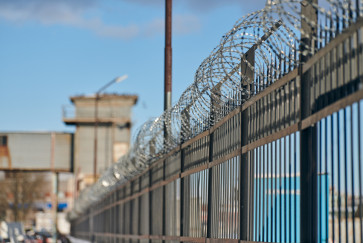 a metal fence topped with barbed wire, with a building and watchtower in the background